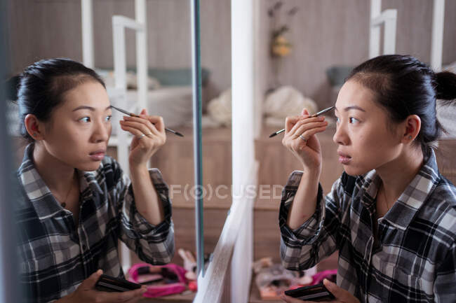 Focalizzato affascinante femmina etnica applicando matita sopracciglio durante il trucco e guardando nello specchio — Foto stock