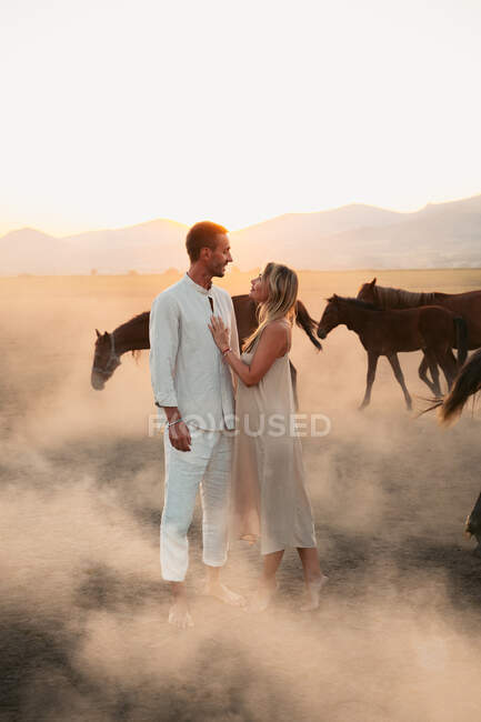 Casal em roupas brancas em pé no campo empoeirado com cavalos e olhando um para o outro em sol na Turquia — Fotografia de Stock