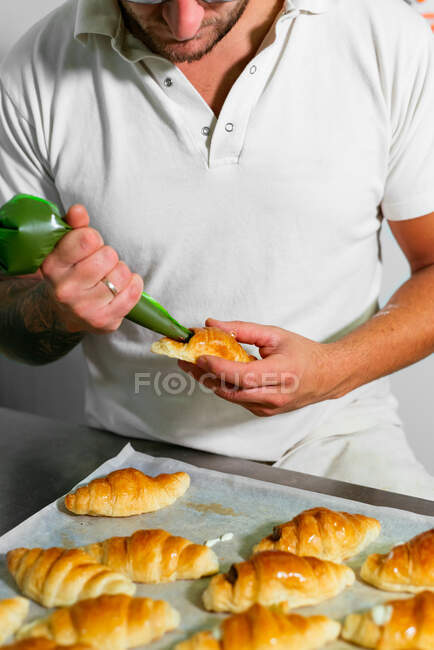 Erntehelfer Bäcker in Freizeitkleidung steht und füllt gebackene Croissants mit Schokoladencreme aus Teigtasche, während er in der Bäckerei arbeitet — Stockfoto