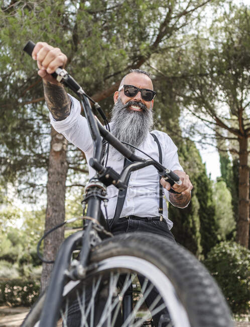 Confiante hipster macho feliz com tatuagens em camisa branca e óculos de sol sentado em bicicleta no parque com árvores verdes na cidade — Fotografia de Stock
