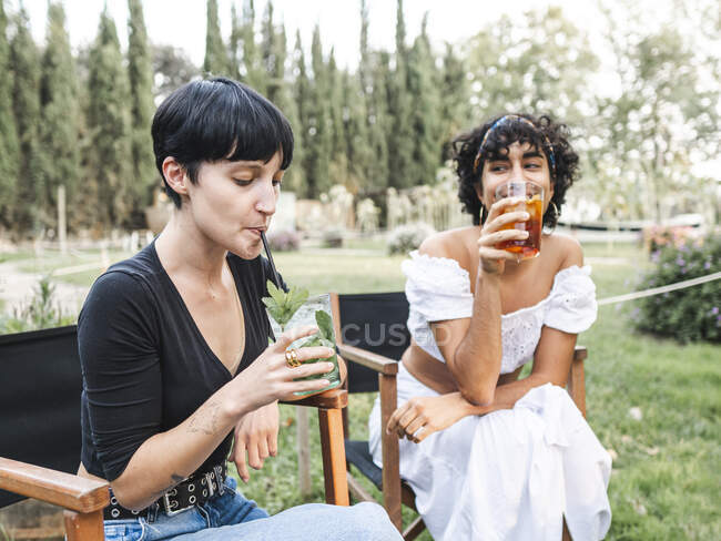 Разнообразные подруги сидят на стульях в летнем парке и пьют освежающий коктейль, наслаждаясь летним днем вместе — стоковое фото