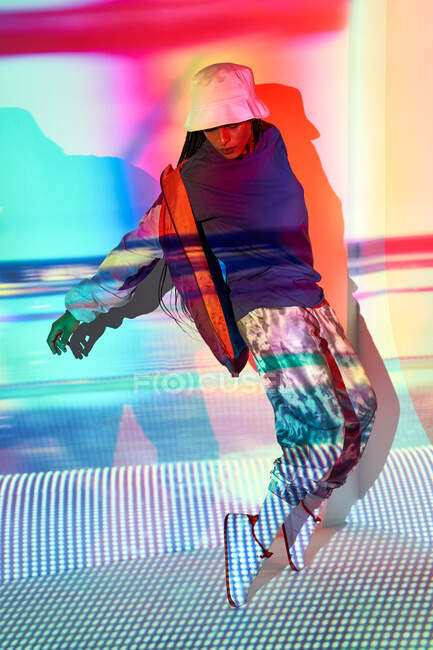 Corpo cheio de confiante jovem étnico feminino millennial em roupa elegante e chapéu de pé em dedos do pé perto da parede com projeção colorida e olhando para baixo — Fotografia de Stock