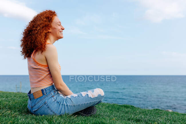 Vista lateral de la sonriente hembra pensativa con el pelo largo de jengibre y los ojos cerrados sentado con las piernas cruzadas en la colina cubierta de hierba en la orilla del mar - foto de stock