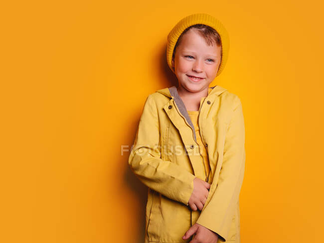 Joyeux garçon veste imperméable jaune vif et bonnet chapeau riant et regardant loin sur fond jaune en studio — Photo de stock