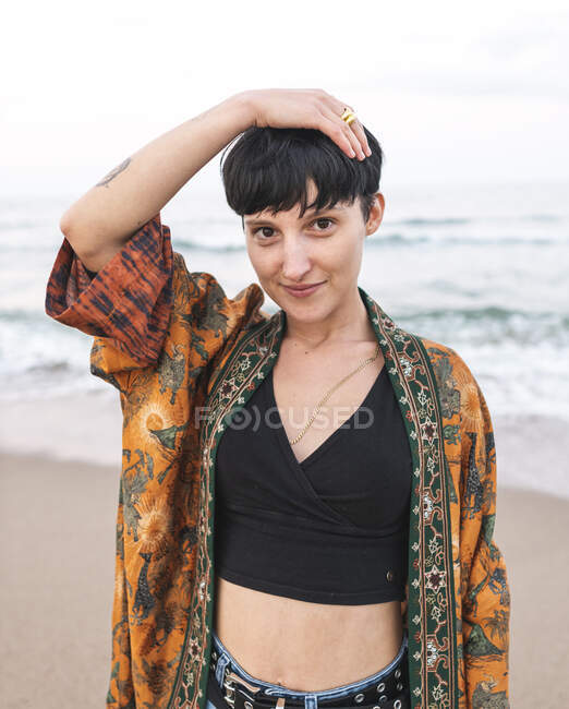 Улыбающаяся женщина в стильной одежде касается головы, стоя на пляже у моря и заходя в камеру — стоковое фото