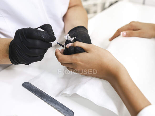 Побочный обзор анонимной маникюрши в перчатках, наносящей лак на ногти молодого клиента контента за столом с инструментами — стоковое фото
