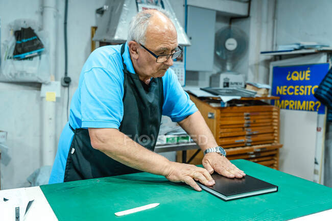 Artesano varón de mediana edad en delantal haciendo encuadernación para libro de pie en la mesa en taller de impresión - foto de stock