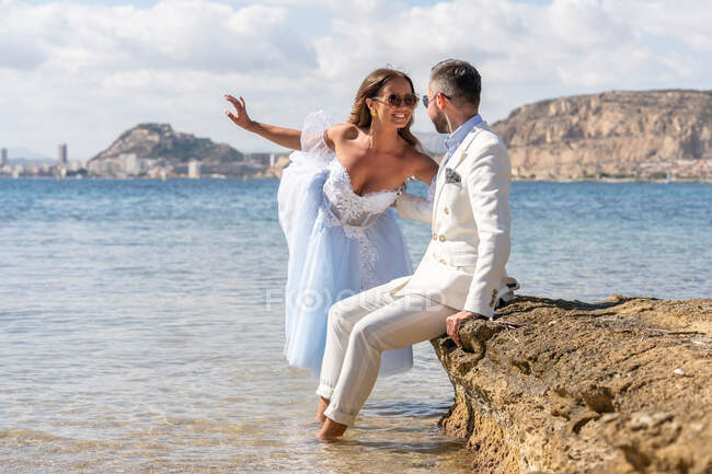 Веселая невеста и жених в элегантных нарядах и солнцезащитных очках веселятся, проводя время в море во время свадебного торжества на природе — стоковое фото