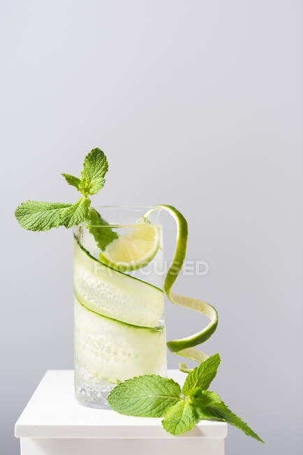 Vaso de refrescante gin tonic con pepino y lima decorado con hojas de menta colocadas sobre mesa blanca sobre fondo gris - foto de stock