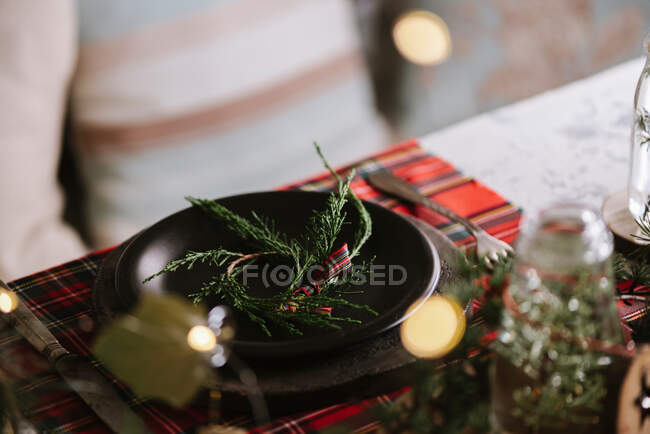 Christmas table setting with wreath on ceramic plate on red checkered tablecloth on the background with lights — Stock Photo