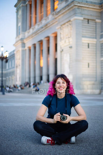 Ganzkörper lächelnde Fotografin mit rosa Haaren und Fotokamera in der Hand, während sie auf einem Gehweg in der Nähe eines alten Gebäudes in der Stadt sitzt — Stockfoto