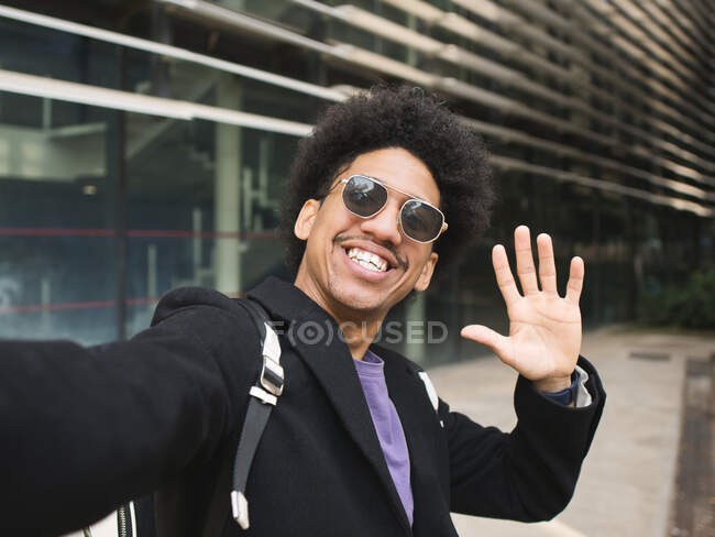 Очарованный молодой афроамериканец в модной одежде и солнцезащитных очках, улыбающийся и машущий рукой, делая селфи на фоне современного коммерческого здания — стоковое фото