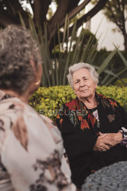Vieille femme souriante assise à table dans un jardin exotique avec des paumes et des buissons et profitant d'une conversation avec une amie âgée — Photo de stock