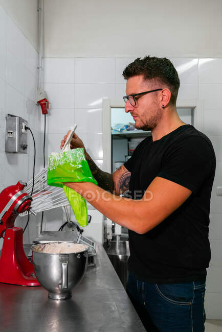 Side view of concentrated young tattooed male baker in casual clothes and eyeglasses filling pastry bag with cream while preparing delicious cake in kitchen — Stock Photo