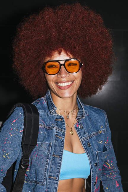 Mujer sonriente con peinado afro y atuendo de moda y gafas de sol mirando a la cámara mientras está de pie sobre fondo negro con mochila en tiempo de noche - foto de stock