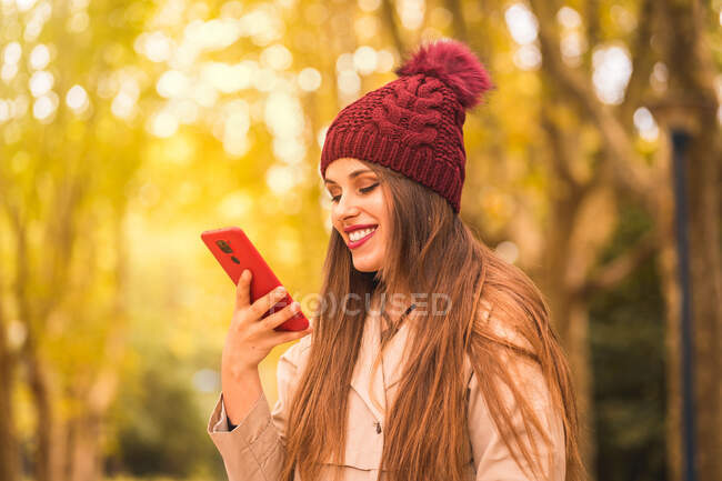 Позитивна жінка з довгим волоссям у зовнішньому одязі із задоволенням посміхається під час смс на мобільному телефоні в парку восени. — стокове фото