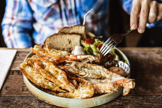 Crop unrecognizable male in casual clothes sitting with fork in hand at wooden table while eating typical Icelandic dish with king prawns and bread in restaurant in daylight — Stock Photo