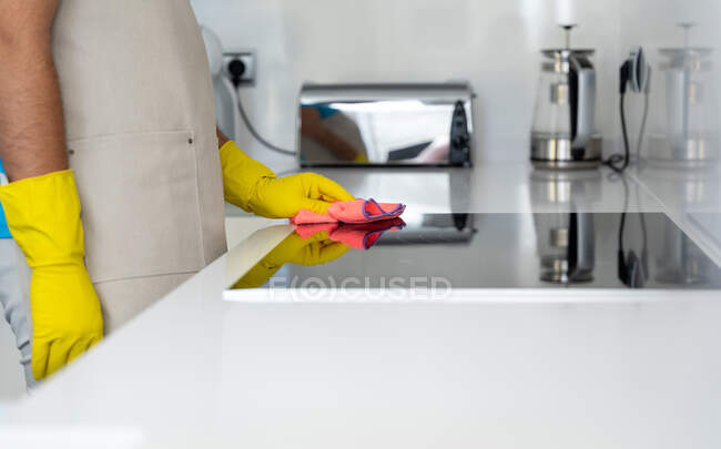 Cosecha irreconocible trabajador masculino en delantal y guantes de goma amarilla limpieza construida en estufa eléctrica con tela en la cocina moderna - foto de stock
