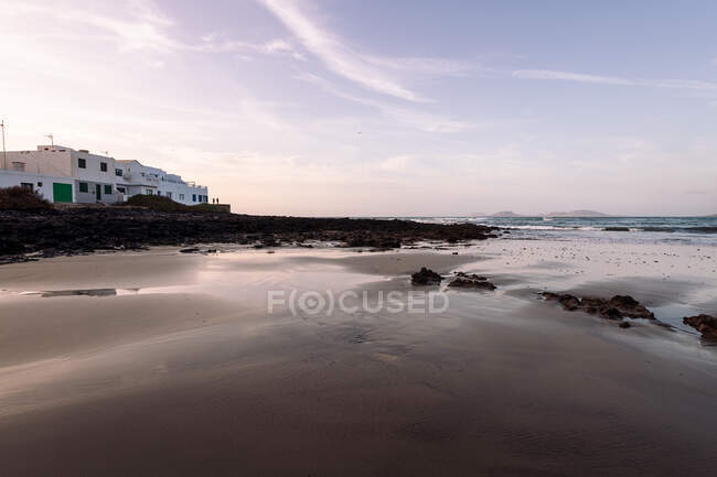 Famara Playa contra el océano con montañas y casa exterior bajo cielo nublado en Teguise Lanzarote Islas Canarias España - foto de stock