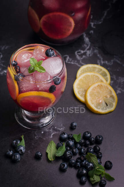 High angle of glass and jug with refreshing cold lemonade with fresh blueberries and lemon slices placed on dark table — Stock Photo