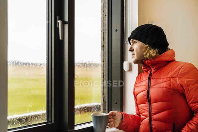 Calme jeune voyageur masculin en veste rouge chaude et chapeau boire une tasse de café chaud et regardant par la fenêtre debout dans la cuisine dans la maison le jour de pluie dans la campagne — Photo de stock