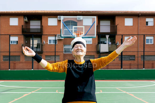 Веселая взрослая женщина с закрытыми глазами в спортивной одежде, стоящая на спортивной площадке с распростёртыми руками возле баскетбольного кольца во время тренировки — стоковое фото