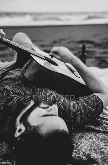 Урожай неузнаваемый мужчина-музыкант в повседневной одежде лежит на песчаном побережье возле размахивая океаном и играть на гитаре при дневном свете — стоковое фото