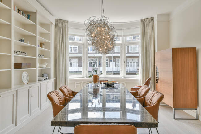 Longa mesa e cadeiras de couro localizadas perto de prateleiras com decorações e janela com cortinas em espaçosa sala de jantar — Fotografia de Stock