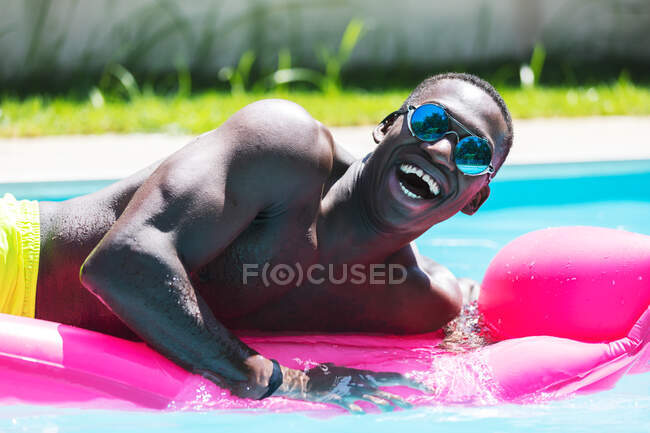 Мужчина без рубашки, афроамериканец в солнечных очках, отдыхает на розовом надувном матрасе в бассейне, загорает в солнечный летний день — стоковое фото