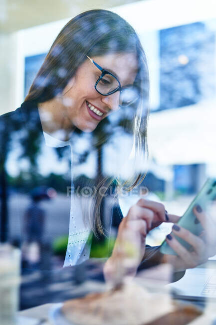 Através de vidro de cultura positiva empregada feminina em óculos usando telefone celular para verificar mensagens de trabalho — Fotografia de Stock