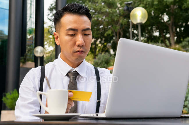 Grave jovem empresário étnico masculino com cartão de crédito sentado na mesa de cafeteria urbana com netbook e xícara de café enquanto faz compras on-line — Fotografia de Stock
