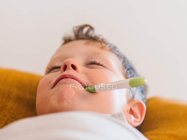 De baixo menino doente medir a temperatura com termômetro eletrônico enquanto deitado no sofá em casa e com gripe — Fotografia de Stock