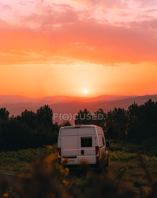 Caravana blanca viajera estacionada en pradera en las tierras altas contra el brillante cielo naranja con sol al atardecer en Galicia - foto de stock