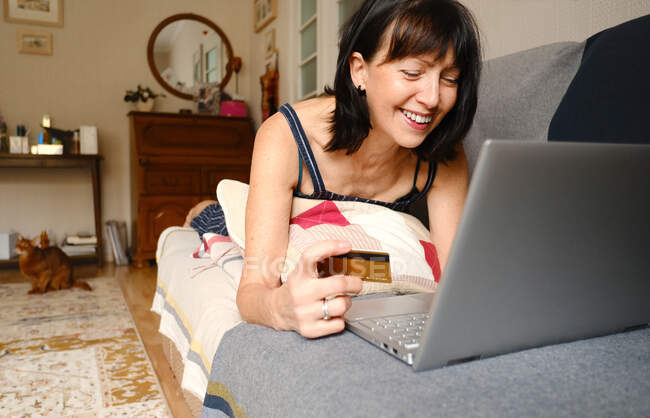Урожай веселый женщина лежит на диване и с помощью пластиковой карты для онлайн оплаты покупок в Интернете на ноутбуке дома — стоковое фото