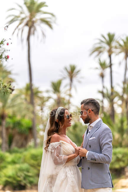 Seitenansicht von glücklichen jungen Eheleuten in Hochzeitskleidern stehend und Händchenhaltend, während sie sich am See und bei Tag im Garten mit grünen Bäumen betrachten — Stockfoto