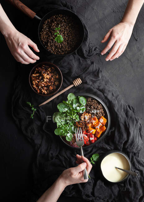 Анонимные люди за столом с чечевицей едят салат с листьями базилика и овощами на черном фоне — стоковое фото