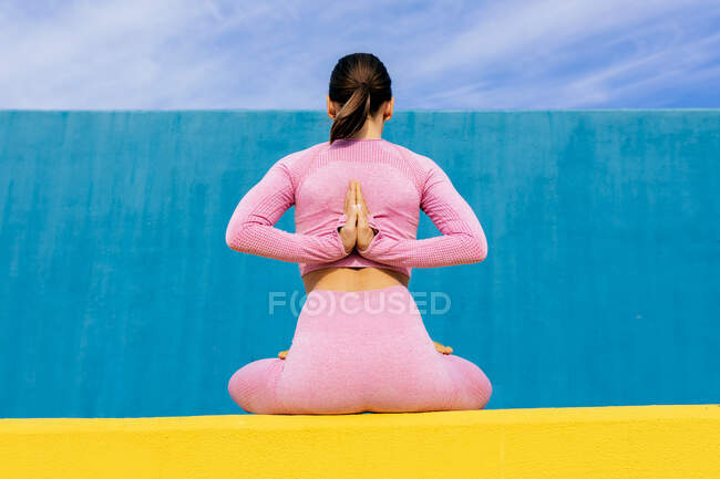 Vista posterior de una hembra delgada con cabello oscuro usando ropa deportiva sentada en la postura de oración inversa con las manos detrás de la espalda y las piernas cruzadas contra la pared azul - foto de stock