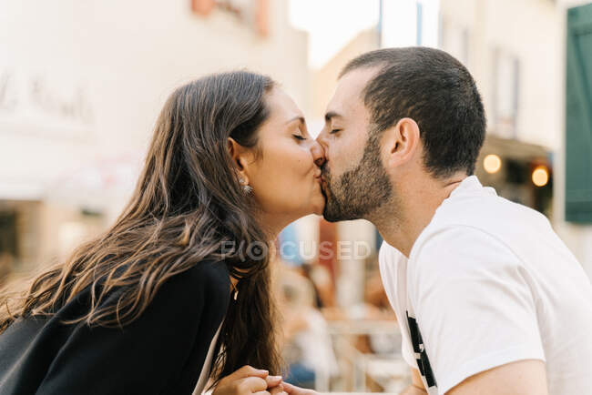 Вид сбоку на позитивную молодую этническую пару целующуюся с закрытыми глазами, сидящую в уличном кафе в солнечный день — стоковое фото