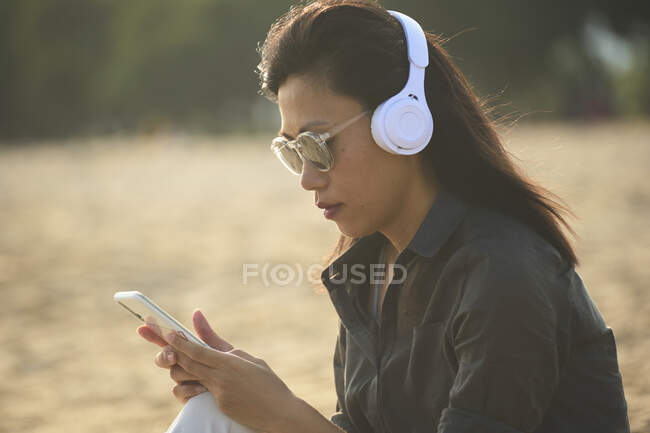 Seitenansicht einer ernsthaften Asiatin mit Sonnenbrille, die SMS auf dem Handy sendet, während sie am Sandstrand über Kopfhörer Musik hört — Stockfoto