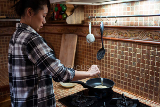 Vista lateral de la joven enfocada en ropa casual cocinando deliciosos panqueques en sartén en la estufa en la cocina - foto de stock