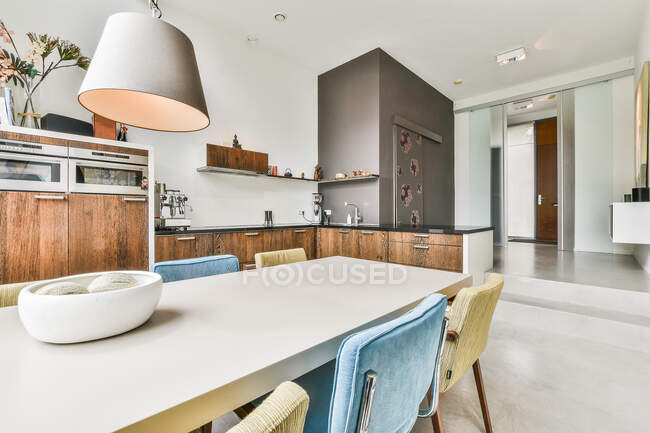Sedie colorate a tavola vicino a armadi in legno con utensili da cucina moderni e mensola in elegante cucina con lampada in appartamento — Foto stock