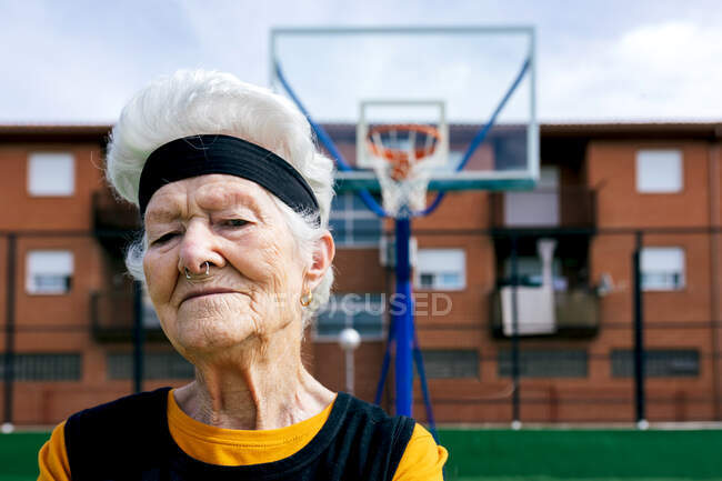 Mujer madura de confianza en ropa deportiva con la nariz perforada mirando a la cámara mientras está de pie en el campo de deportes durante el entrenamiento en la calle - foto de stock