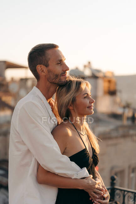 Vista lateral de abraçar homem e mulher apaixonados de pé juntos na rua e olhando para longe contra o pôr do sol — Fotografia de Stock