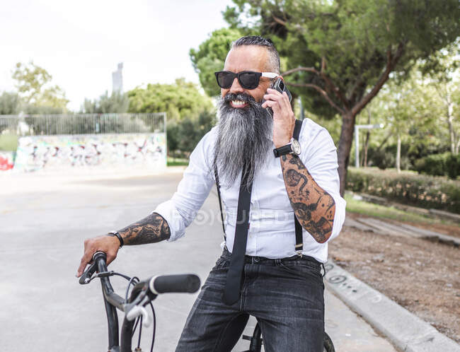 Bearded male in sunglasses and white shirt speaking on smartphone while sitting on bicycle in park with trees — Stock Photo