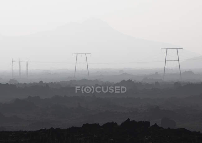 Black and white of power lines with wires placed on ground with rough surface against mountain range covered with fog — Stock Photo