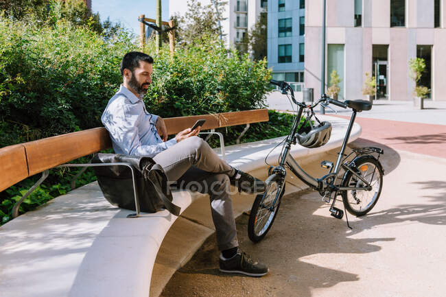 Ganzkörper-Seitenansicht eines männlichen Managers, der auf Bank neben Fahrrad sitzt und im Park Nachrichten auf dem Smartphone liest — Stockfoto