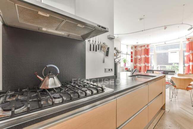 Metal kettle on gas cooker under extractor hood near kitchen cabinets in stylish apartment with window in light living room — Stock Photo