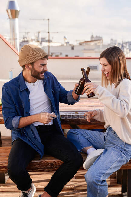 Очарованные юные друзья в повседневной одежде, сидящие на деревянной скамейке и звенящие бутылки пива во время отдыха на террасе вместе — стоковое фото