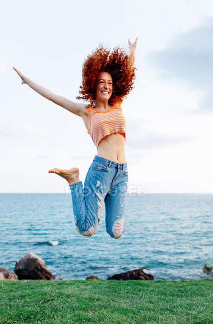 Corpo inteiro de mulher enérgica feliz com cabelo encaracolado de gengibre voador pulando acima do chão gramado na costa do mar ondulante azul olhando para longe — Fotografia de Stock