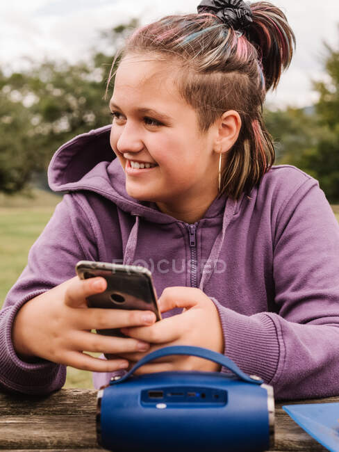 Приємна дівчина - підліток пише повідомлення по мобільному телефону і озирається, сидячи за столом, з переносними промовцями за своєю природою з деревами. — стокове фото